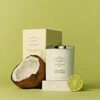 A coconut and lime candle with a silver lid is on a light green background, alongside half a coconut and a slice of lime, conveying a fresh, tropical vibe.