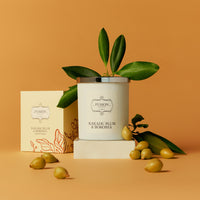 A white candle labeled "Fusion Kakadu Plum and Boronia" with green leaves on top sits beside a matching box on an orange background. Small Kakadu Plums scattered around convey freshness.