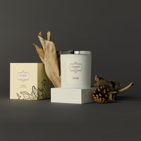 Elegant candle display featuring a white candle with a silver lid beside its box, accented by dried leaves and pinecones, set against a dark backdrop.