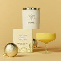 A decorative scene with a candle labelled Brandy Custard, glass with custard in it, and the packaging box against a vibrant yellow backdrop, featuring decorative Christmas Ornaments.