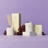 A group photo featuring a white glass jar candle and matching white glass jar diffuser surrounded by decorative dark purple grapes against a vibrant purple background.