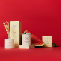 A box of watermelon-scented candles next to a fresh watermelon and lychees, showcasing a vibrant and fruity theme.